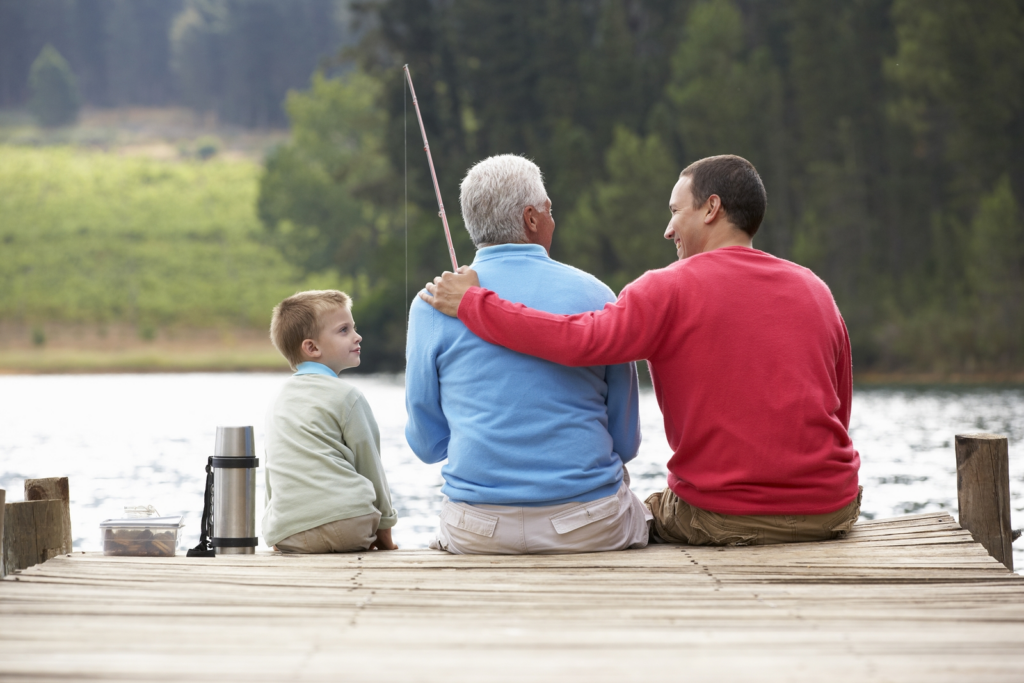 Grandfather, son, and grandson sitting at the edge of a dock smiling and fishing.